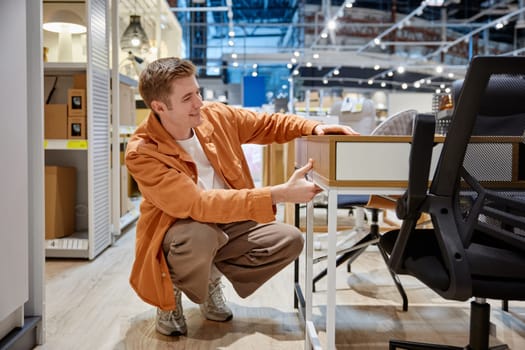Young man choosing desk table for home office workplace interior remodeling. Purchasing with discount at big shopping mall