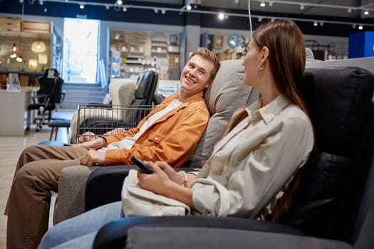 Young man and woman loving couple sitting in comfortable armchairs while choosing one for home stylish interior at furniture shop store department