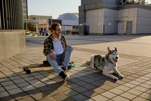 Cheerful young hipster guy using smartphone while resting with dog after skateboarding. Handsome man sitting on longboard shooting selfie for social media