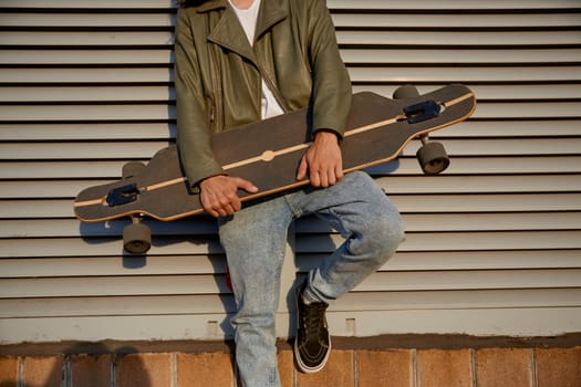 Crop of urban skater man posing for camera with longboard standing in front of street building wall