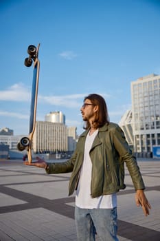 Hipster guy holding skateboard on hand in balance doing trick. Cool teenager man enjoying urban style outdoors