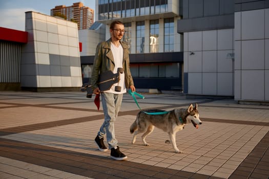 Hipster guy in casual clothing skateboarding and walking dog moving together through city square. Pet owner and loving puppy enjoying physical activity during calm evening at downtown district