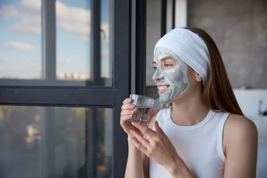 Young attractive woman morning routine for health and beauty. Charming girl with cosmetics facial mask drinking water from glass while dreamily looking in window. Closeup female portrait
