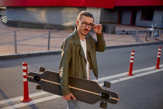 Portrait of handsome casual guy with skateboard over downtown background. Hipster man touching trendy eyeglasses holding longboard in hands. Carefree pastime, sportive leisure lifestyle