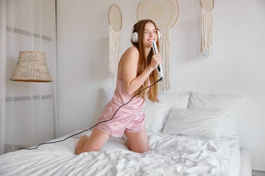 Young pretty woman wearing pajama and headphones listening to favorite music singing like star using electric hair straighter as microphone