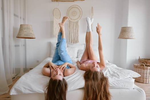 Two young beautiful girl friends wearing pajamas enjoying free time together lying on bed in bedchamber with raised up legs. Friendship and gossiping during slumber party celebration concept