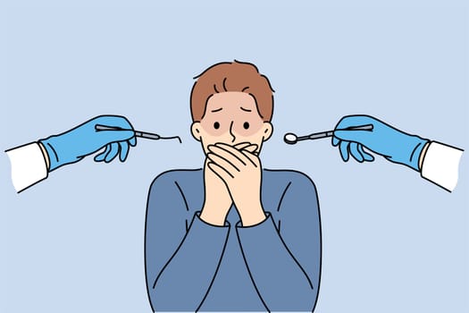 Man suffering from dentophobia closes mouth and stands near hands with dental devices. Guy is afraid of doctors at dental clinic, and feels panic, not allowing caries to be treated