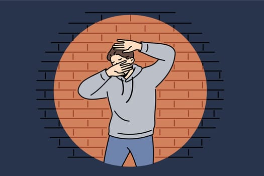 Man criminal in light of police lantern stands near brick wall and covers face with hands. Young guy criminal who committed robbery or murder was cornered after pursuit and investigation