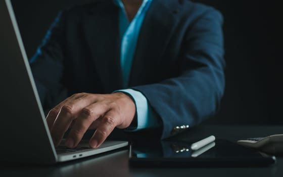 Businessman in black suit working on laptop computer and tablet, Hand typing on keyboard on table at office with dark background, Online working, Close up, Copy space.