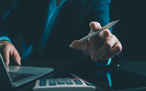 Businessman in black suit working on laptop computer and tablet and calculator, Hand Hands touch on tablet at office with dark background, Online working, Close up, Copy space.