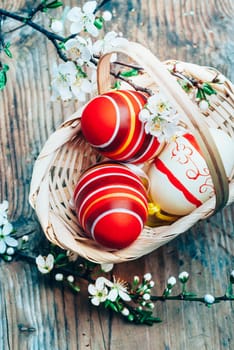 Top view of colorful painted easter eggs in basket and cherry blossom branch decor on vintage wooden table. Traditional seasonal holiday still life from above. Easter celebrate and spring concept.
