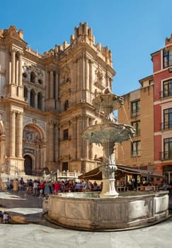 Málaga, Spain - September 28,2023: Facade of the Malaga Cathedral or Santa Iglesia Catedral Basílica de la Encarnación, with a fountain and tourists in the town square in the foreground.