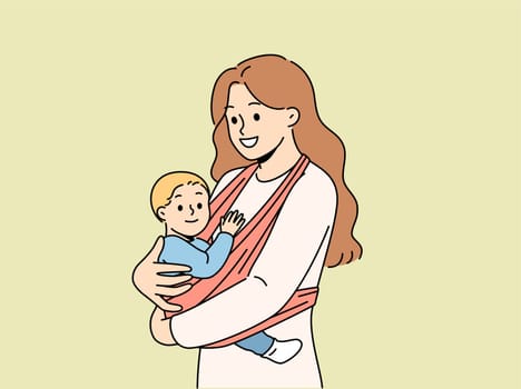 Mother holds baby in carrier sling and smiles, taking care of son and using comfortable babywearing. Caring happy woman with newborn, recommends purchasing sling for young mothers.