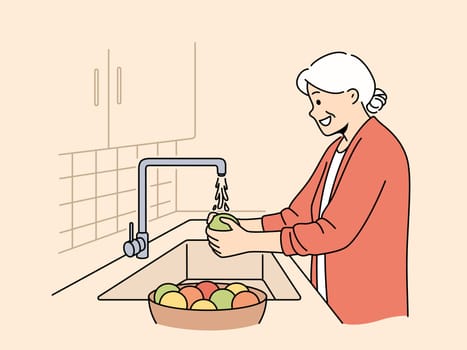 Elderly woman washes fruits in sink, standing in kitchen and observing rules of hygiene to avoid infection. Grandmother washes vegetables before preparing dinner, following recommendations of doctors