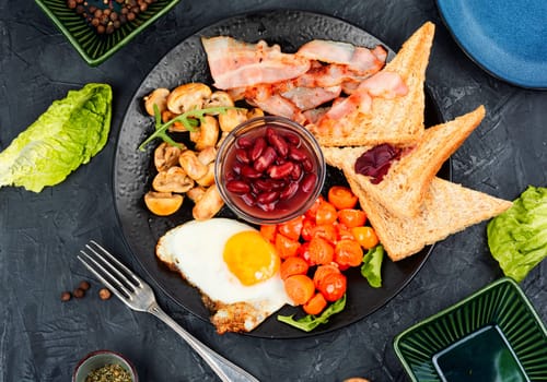 English breakfast: fried egg, fried bacon,vegetables, beans and toast on a plate.