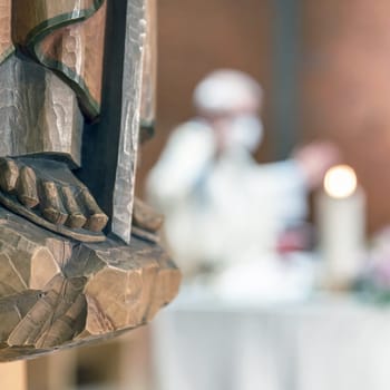 Holy wooden statue. Carved wooden feet, detail of wooden statue of a Saint. In the blurred background a priest celebrates Mass.