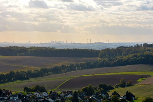 Top view of a green landscape with houses of a village, farmland, forest and blue sky. In the background the skyline of the city of Frankfurt am Main, Hesse, Germany