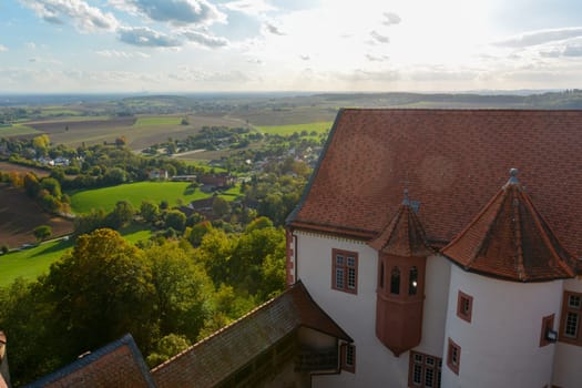 Top view of the roof of an old castle and a green landscape with houses of a village, farmland, forest and blue sky