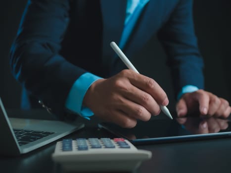 Businessman in black suit working on laptop computer and tablet and calculator, Hand Hands touch on tablet at office with dark background, Online working, Close up, Copy space.
