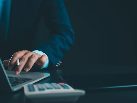 Businessman in black suit working on laptop computer and tablet and calculator, Hand Hands touch on tablet at office with dark background, Online working, Close up, Copy space.