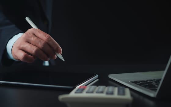 Businessman in black suit working on laptop computer and tablet and calculator, Hand Hands touch on tablet at office with dark background, Online working, Close up, Copy space.