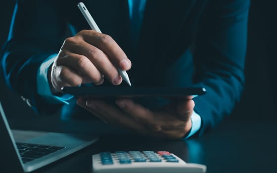 Businessman in black suit working on laptop computer and tablet and calculator, Hand Hands touch on tablet at office with dark background, Online working, Close up, Copy space.