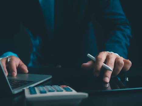 Businessman in black suit working on laptop computer and tablet and calculator, Hand Hands touch on tablet at office with dark background, Online working, Close up, Copy space.
