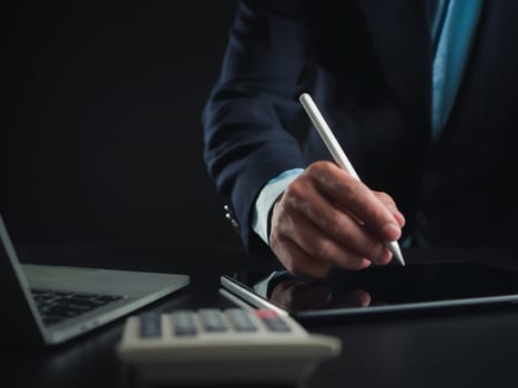 Businessman in black suit working on laptop computer and tablet and calculator, Hand Hands touch on tablet at office with dark background, Online working, Close up, Copy space.