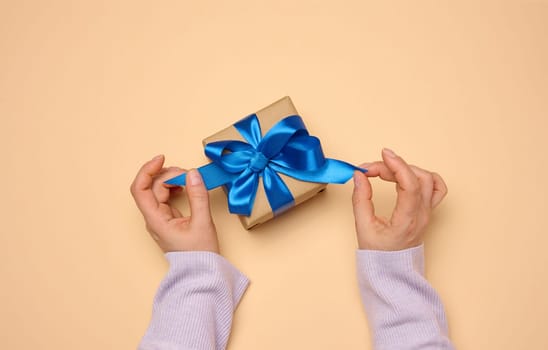 A woman's hand holds a gift box wrapped in a blue silk ribbon on a beige background, top view