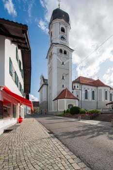 North-side wide angle view of the St. Andreas Church with bell tower in Nesselwang / Bavaria from in front of a blue sky with clouds and lens flares in the summer of 2023.