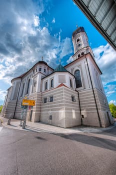 Extreme wide angle view of the southeast corner of the Saint Andreas Church in Nesselwang Bavaria with a view over the road junction to Markoberdorf against a blue sky with clouds in summer 2023.