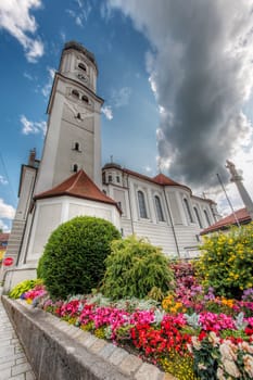 North-side extreme wide-angle view of the Saint Andreas Church with the bell tower and flower beds in front of it in Nesselwang/Bavaria against a blue sky with clouds in summer 2023.