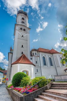 North-side wide-angle view of the St. Andrew's Church with the bell tower and flower beds in front of it in Nesselwang/Bavaria against a blue sky with clouds in the summer of 2023.