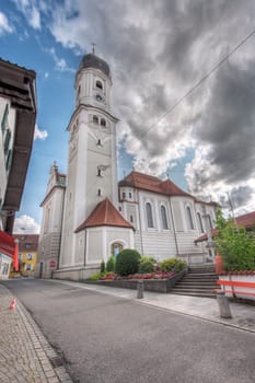 North-side wide angle view of the St. Andreas Church with bell tower in Nesselwang / Bavaria from in front of a blue sky with clouds and lens flares in the summer of 2023.