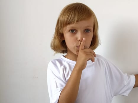 Silence gesture. The finger is near the mouth. The girl on a white background shows a gesture of keeping silence. Child of 7 years old with blond hair. Caucasian type. Copy space. Place for text