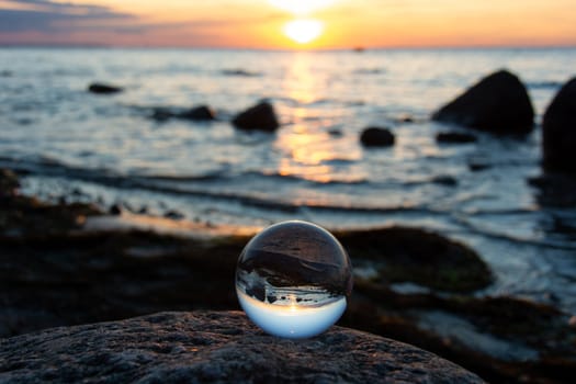 Glass ball on a rock at sunset on the beach, the sea and the setting sun are reflected in the ball