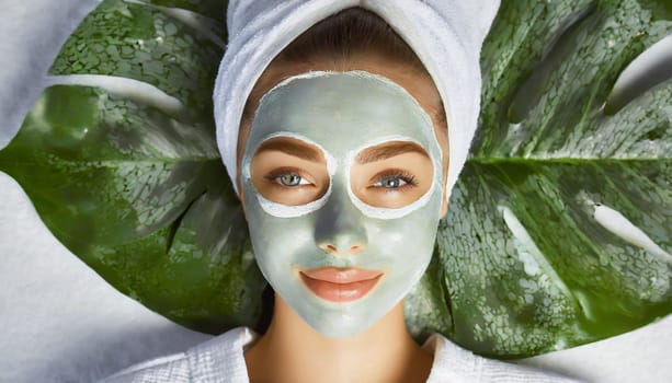 Skin care. Cheerful attractive caucasian female with clay mask on face and cucumber slices in hands against blue background getting beauty treatment. Spa concept with copy space