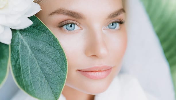 Portrait of beautiful woman with healthy glow perfect smooth skin holds tropical leaf, look into the hole of green paper. Young girl enjoys cream facial mask. Fashion, beauty, skincare concept.
