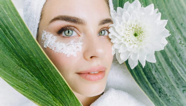 Portrait of beautiful woman with healthy glow perfect smooth skin holds tropical leaf, look into the hole of green paper. Young girl enjoys cream facial mask. Fashion, beauty, skincare concept.