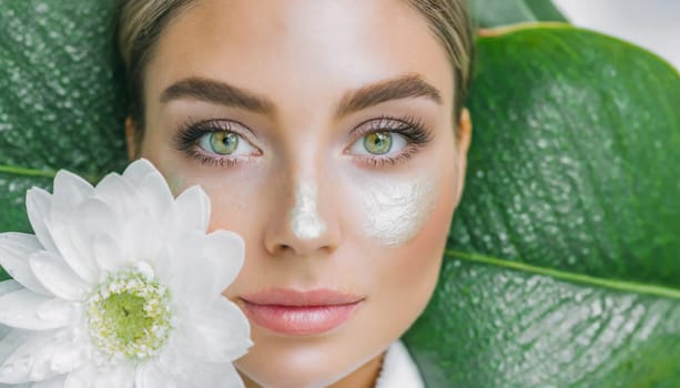 Portrait of beautiful woman with healthy glow perfect smooth skin holds tropical leaf, look into the hole of green paper. Young girl enjoys cream facial mask. Fashion, beauty, skincare concept.