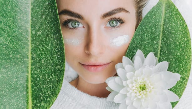Portrait of beautiful woman with healthy glow perfect smooth skin holds tropical leaf, look into the hole of green paper. Young girl enjoys cream facial mask. Fashion, beauty, skincare concept.