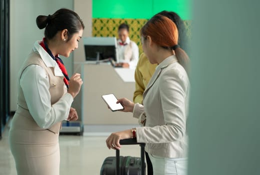 Young businesswoman using smartphone while standing with luggage in airport check in terminal. Passenger showing boarding document on phone with a ground attendant