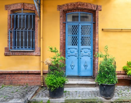 Charming blue metal door adorned with a vintage knob and wrought iron window adding a pop of color to a warm yellow building. Lush greenery spills from plant pots lining a cobblestone street