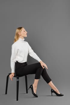 Young woman in tight trousers and white sweater sits in profile in studio