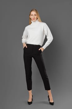 Slender tall blonde woman in tight breeches and a sweater posing in the studio on a grey background
