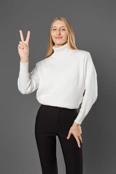Young woman in trousers and sweater shows victory sign lifting two fingers up