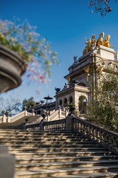 A view of the side staircase of the Cascada Monumental in the Ciutadella Park in Barcelona, Spain. Above the arch is a giant gilded iron sculpture representing an allegory of dawn (Aurora in Latin), standing on a chariot drawn by four horses: the Quadriga of the Aurora.