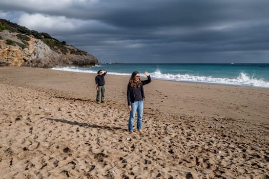 Two women standing on a beach, making the same gesture to protect their eyes from the sunlight. Young mother and daughter protect their eyes with their hand and look in the same direction. Overcast sky, rough sea. Copy space for a text.