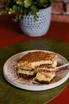 Close-up of a portion of Tiramisu Cake with cocoa powder dusting served with a fork on a white plate. Green plant in a vase on the restaurant table, blurred background.