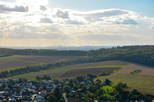 Top view of a green landscape with houses of a village, farmland, forest and blue sky. In the background the skyline of the city of Frankfurt am Main, Hesse, Germany
